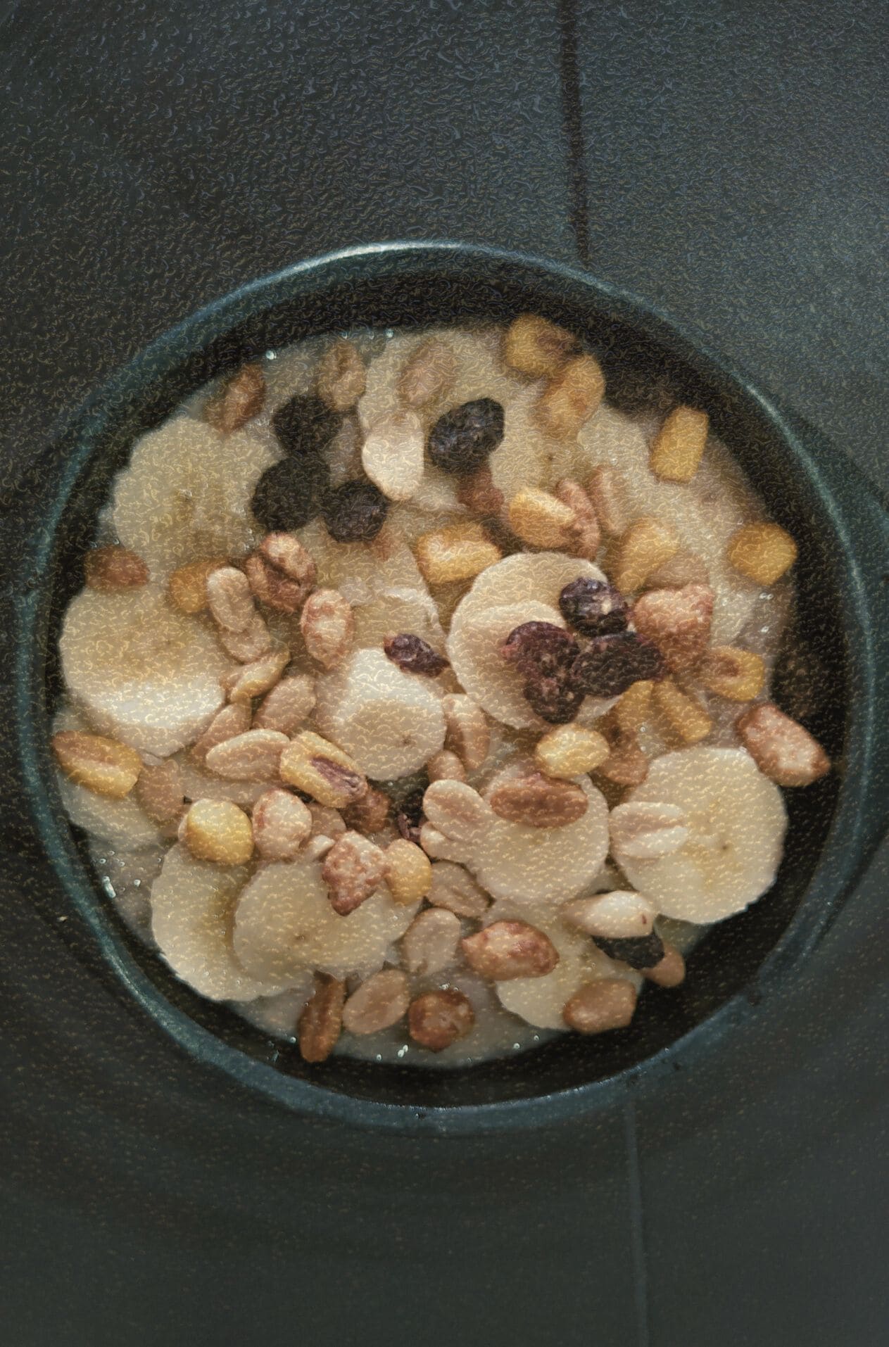 Fotografía cenital de un bowl oscuro con avena, rodajas de banano, pasas y nueces mixtas. Un bodegón cinematográfico que documenta la alimentación real y los momentos de mesa cotidianos en el archivo KOKOGOA.