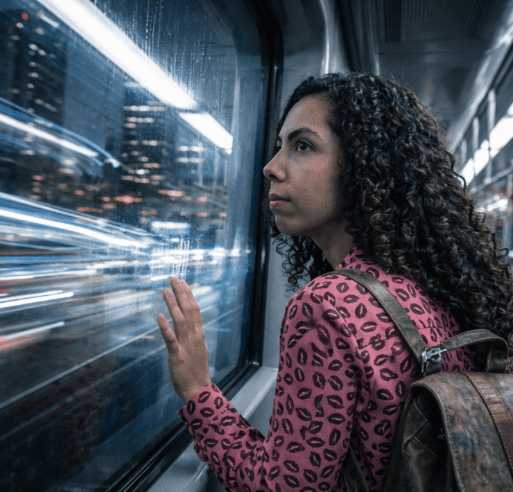 Anlli en el Metro de Medellín: Retrato detallado de una mujer joven con cabello rizado apoyada en la ventana del metro de Medellín; las ráfagas de luz nocturna y el rastro de movimiento al fondo reflejan la velocidad del transporte urbano, en una jornada de trabajo que empieza en la madrugada.