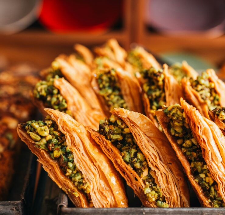 Baklava en Estambul: Foto en primer plano de filas de baklava turca dispuestas en una bandeja metálica de horno. Se aprecian las finas capas de masa filo en color dorado crujiente y el intenso relleno de pistacho picado.