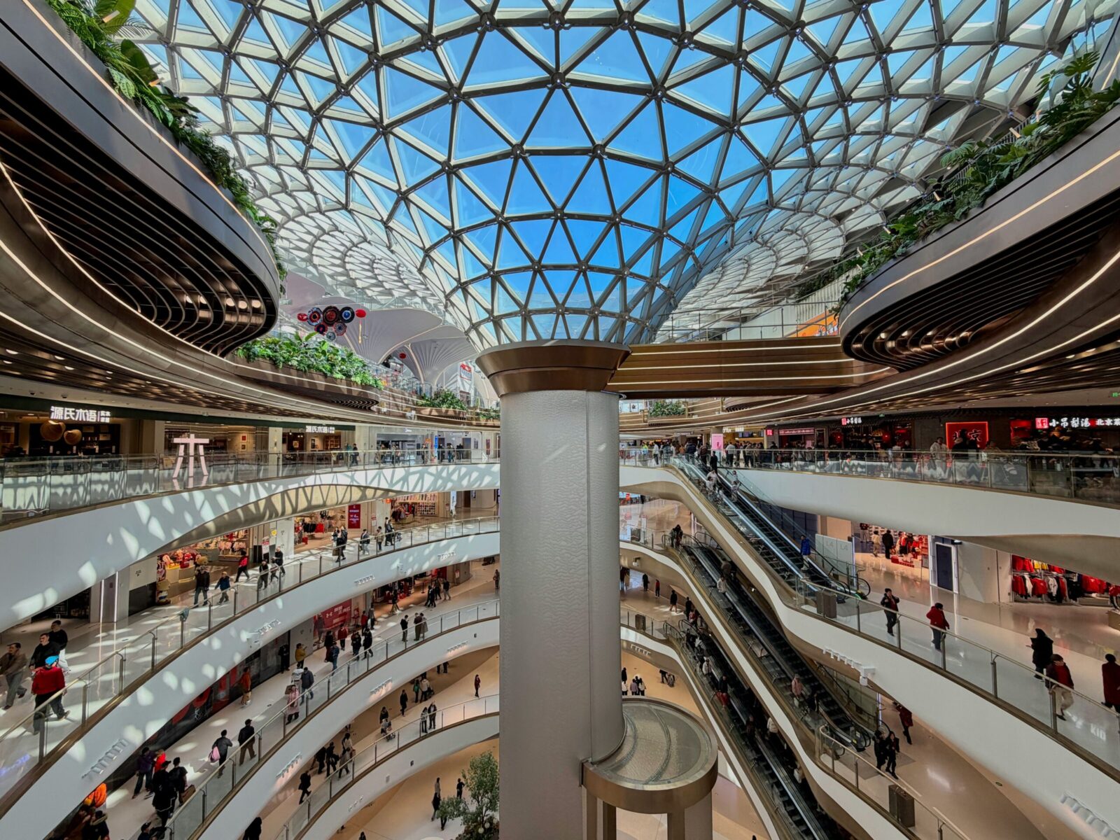 Sociedad de consumo: vista interior de un centro comercial futurista y ultra-moderno con múltiples niveles curvos de mármol blanco y una imponente cúpula de vidrio geométrica. La imagen captura la escala monumental de un ecosistema climatizado donde pequeñas figuras humanas circulan por pasillos y escaleras mecánicas bajo una luz blanca y uniforme.