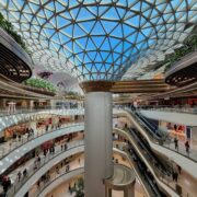 Sociedad de consumo: vista interior de un centro comercial futurista y ultra-moderno con múltiples niveles curvos de mármol blanco y una imponente cúpula de vidrio geométrica. La imagen captura la escala monumental de un ecosistema climatizado donde pequeñas figuras humanas circulan por pasillos y escaleras mecánicas bajo una luz blanca y uniforme.