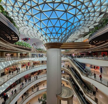 Sociedad de consumo: vista interior de un centro comercial futurista y ultra-moderno con múltiples niveles curvos de mármol blanco y una imponente cúpula de vidrio geométrica. La imagen captura la escala monumental de un ecosistema climatizado donde pequeñas figuras humanas circulan por pasillos y escaleras mecánicas bajo una luz blanca y uniforme.
