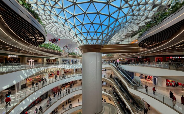 Sociedad de consumo: vista interior de un centro comercial futurista y ultra-moderno con múltiples niveles curvos de mármol blanco y una imponente cúpula de vidrio geométrica. La imagen captura la escala monumental de un ecosistema climatizado donde pequeñas figuras humanas circulan por pasillos y escaleras mecánicas bajo una luz blanca y uniforme.