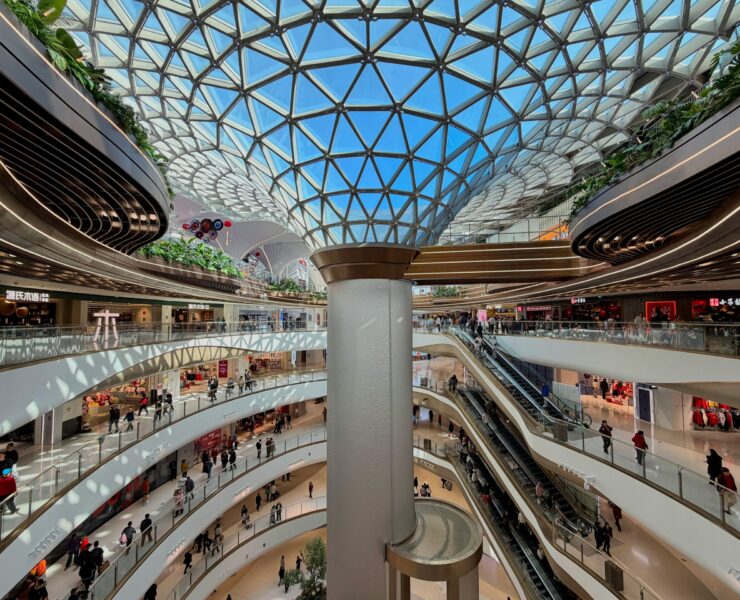 Sociedad de consumo: vista interior de un centro comercial futurista y ultra-moderno con múltiples niveles curvos de mármol blanco y una imponente cúpula de vidrio geométrica. La imagen captura la escala monumental de un ecosistema climatizado donde pequeñas figuras humanas circulan por pasillos y escaleras mecánicas bajo una luz blanca y uniforme.