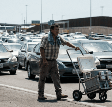 Manuel Antonio en la Garita de san Ysidro: Imagen de un hombre con un trapo azul rey sobre los hombros mientras empuja un carrito metálico de refrescos. Al fondo, una fila interminable de vehículos transita la frontera entre Tijuana y San Diego bajo un sol intenso.