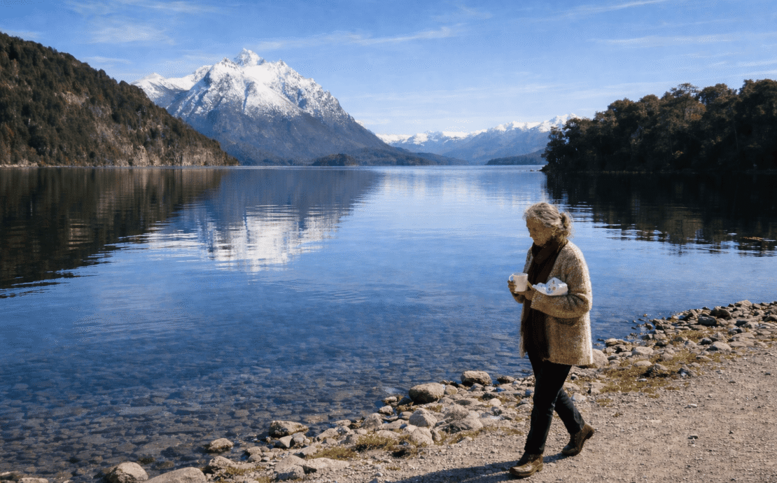 María Soledad en Bariloche: Imagen de una mujer de 75 años caminando por la orilla del lago Nahuel Huapi en Bariloche. Viste un saco de lana artesanal, sostiene una taza de cerámica y un paquete de chocolates, con montañas nevadas y agua cristalina de fondo.