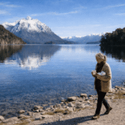 María Soledad en Bariloche: Imagen de una mujer de 75 años caminando por la orilla del lago Nahuel Huapi en Bariloche. Viste un saco de lana artesanal, sostiene una taza de cerámica y un paquete de chocolates, con montañas nevadas y agua cristalina de fondo.