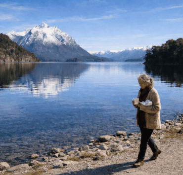 María Soledad en Bariloche: Imagen de una mujer de 75 años caminando por la orilla del lago Nahuel Huapi en Bariloche. Viste un saco de lana artesanal, sostiene una taza de cerámica y un paquete de chocolates, con montañas nevadas y agua cristalina de fondo.