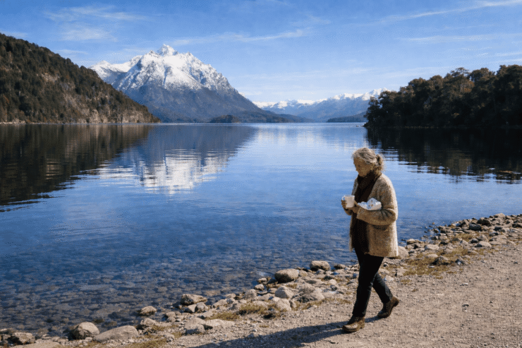 María Soledad en Bariloche: Imagen de una mujer de 75 años caminando por la orilla del lago Nahuel Huapi en Bariloche. Viste un saco de lana artesanal, sostiene una taza de cerámica y un paquete de chocolates, con montañas nevadas y agua cristalina de fondo.