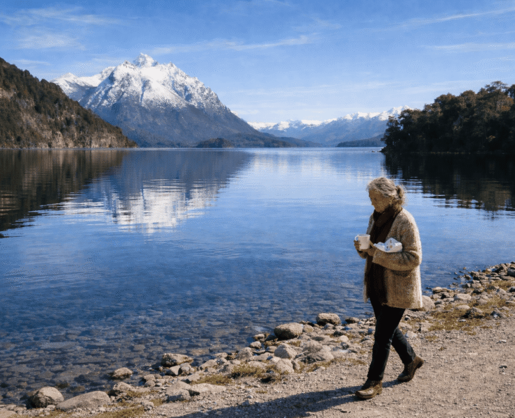 María Soledad en Bariloche: Imagen de una mujer de 75 años caminando por la orilla del lago Nahuel Huapi en Bariloche. Viste un saco de lana artesanal, sostiene una taza de cerámica y un paquete de chocolates, con montañas nevadas y agua cristalina de fondo.