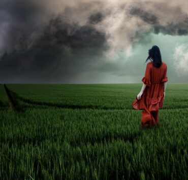 Fotografía de una mujer de espaldas con un vestido rojo largo, caminando sola por un inmenso campo de trigo verde. Al fondo, un cielo dramático cubierto de nubes negras de tormenta. La imagen evoca una profunda sensación de soledad y desamparo emocional ante la incertidumbre del horizonte.