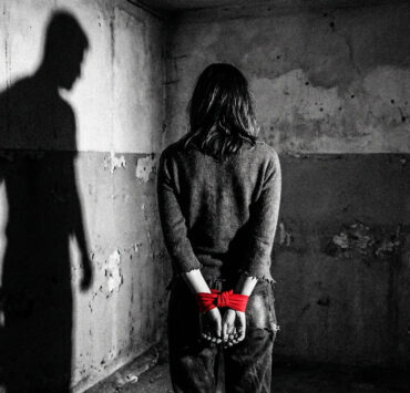 Fotografía conceptual en blanco y negro de una mujer de espaldas, con las manos atadas por una cinta roja vibrante, en una habitación de hormigón desgastado. A su izquierda, se proyecta una sombra masculina amenazante sobre la pared. Una metáfora visual de la violencia patriarcal y el miedo habitado.