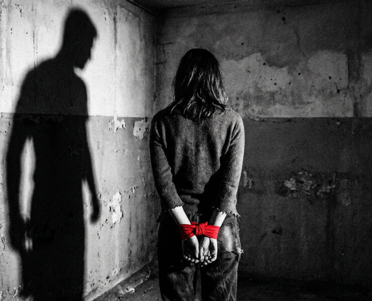 Fotografía conceptual en blanco y negro de una mujer de espaldas, con las manos atadas por una cinta roja vibrante, en una habitación de hormigón desgastado. A su izquierda, se proyecta una sombra masculina amenazante sobre la pared. Una metáfora visual de la violencia patriarcal y el miedo habitado.