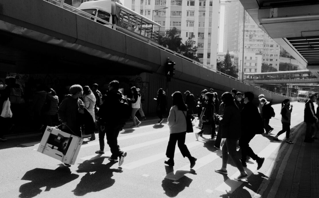 Fotografía de calle en blanco y negro con un marcado contraste lumínico. Una masa de transeúntes cruza una avenida bajo la sombra de una estructura elevada; las siluetas oscuras se recortan contra el asfalto brillante, destacando la inercia del movimiento colectivo en la ciudad.