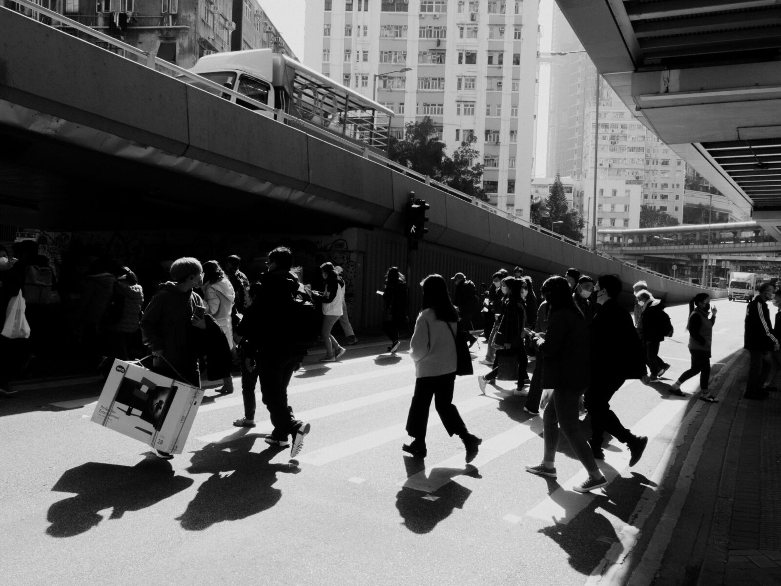 Fotografía de calle en blanco y negro con un marcado contraste lumínico. Una masa de transeúntes cruza una avenida bajo la sombra de una estructura elevada; las siluetas oscuras se recortan contra el asfalto brillante, destacando la inercia del movimiento colectivo en la ciudad.