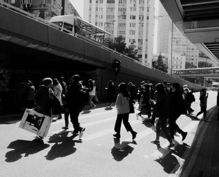 Fotografía de calle en blanco y negro con un marcado contraste lumínico. Una masa de transeúntes cruza una avenida bajo la sombra de una estructura elevada; las siluetas oscuras se recortan contra el asfalto brillante, destacando la inercia del movimiento colectivo en la ciudad.