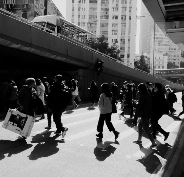 Fotografía de calle en blanco y negro con un marcado contraste lumínico. Una masa de transeúntes cruza una avenida bajo la sombra de una estructura elevada; las siluetas oscuras se recortan contra el asfalto brillante, destacando la inercia del movimiento colectivo en la ciudad.