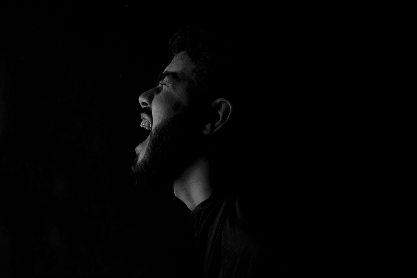 Fotografía en blanco y negro de alto contraste que muestra el perfil de un hombre con barba gritando con fuerza hacia la oscuridad. La iluminación lateral resalta la expresión de angustia y desahogo sobre un fondo completamente negro. Una representación visual del dolor y el vacío que deja un amor imaginario.