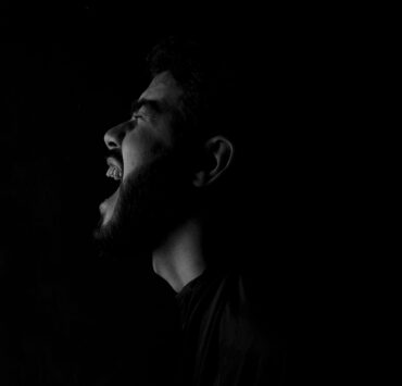 Fotografía en blanco y negro de alto contraste que muestra el perfil de un hombre con barba gritando con fuerza hacia la oscuridad. La iluminación lateral resalta la expresión de angustia y desahogo sobre un fondo completamente negro. Una representación visual del dolor y el vacío que deja un amor imaginario.