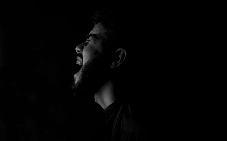 Fotografía en blanco y negro de alto contraste que muestra el perfil de un hombre con barba gritando con fuerza hacia la oscuridad. La iluminación lateral resalta la expresión de angustia y desahogo sobre un fondo completamente negro. Una representación visual del dolor y el vacío que deja un amor imaginario.