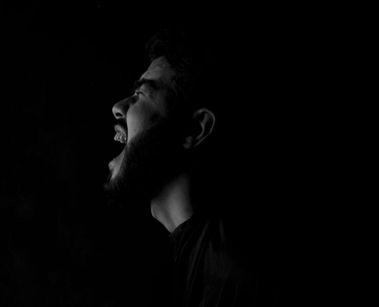 Fotografía en blanco y negro de alto contraste que muestra el perfil de un hombre con barba gritando con fuerza hacia la oscuridad. La iluminación lateral resalta la expresión de angustia y desahogo sobre un fondo completamente negro. Una representación visual del dolor y el vacío que deja un amor imaginario.