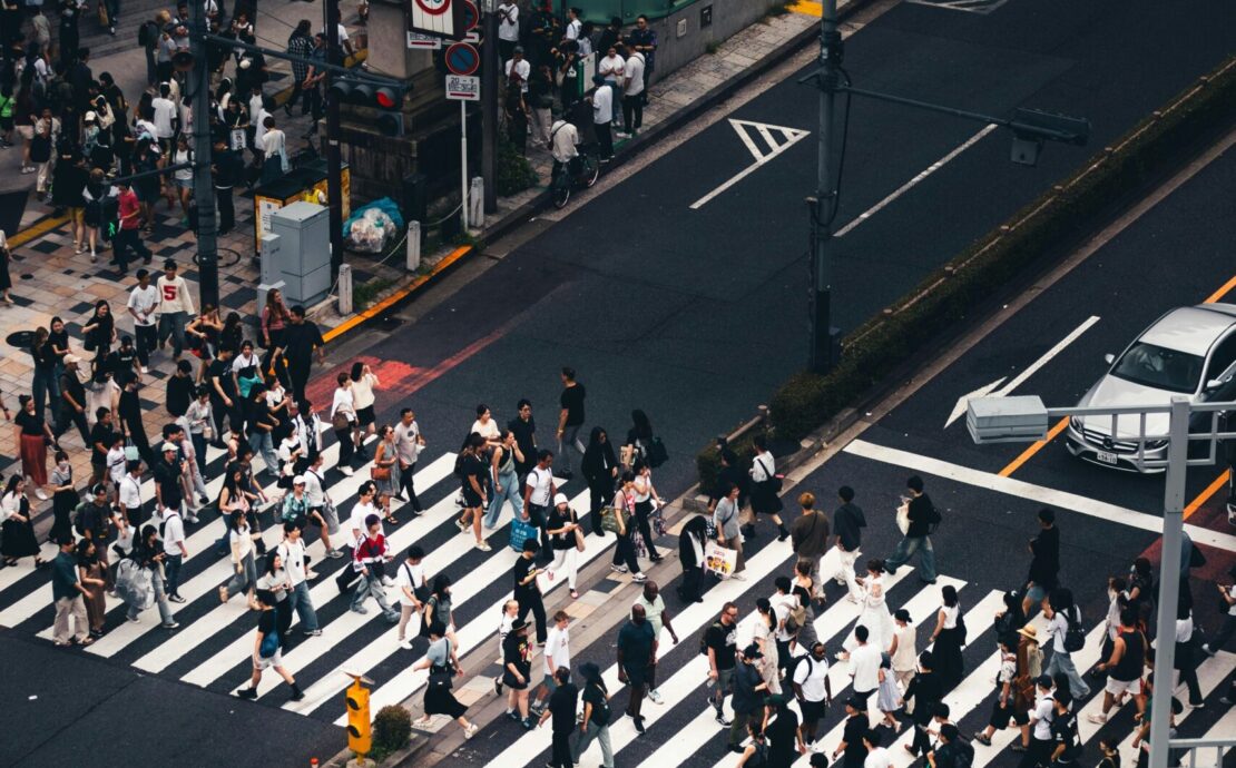 Soledad hiperconectada: Fotografía cenital de alto contraste sobre un cruce peatonal. Una marea humana de cientos de transeúntes, reducidos a siluetas desde las alturas, atraviesa de forma asincrónica las gruesas franjas blancas de un paso de cebra. La composición resalta el asfalto gris oscuro, vehículos de colores sobrios detenidos y la señalética vial, creando una retícula mecánica donde el individuo se disuelve en un flujo de movimiento masivo y geométrico.