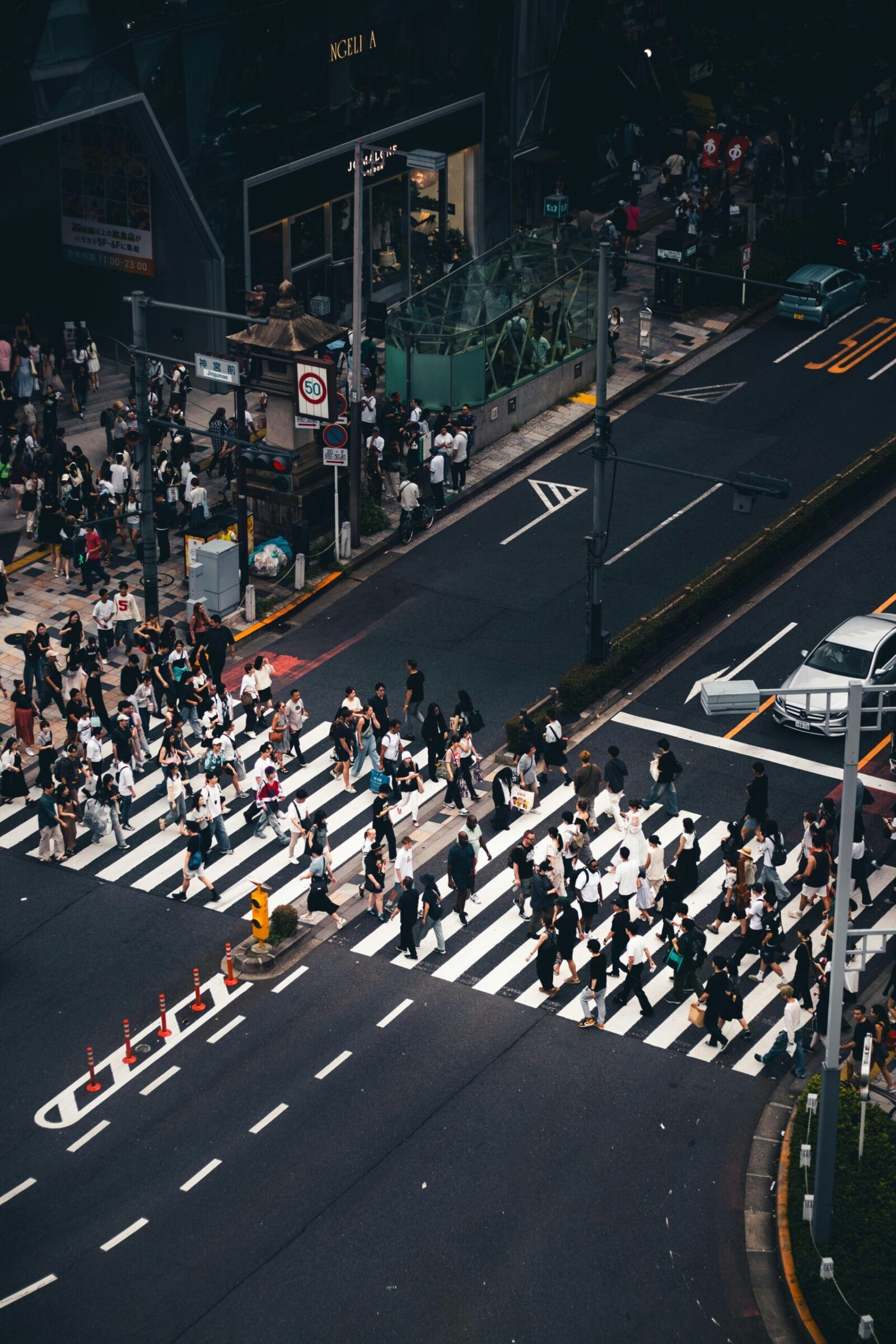 Soledad hiperconectada: Fotografía cenital de alto contraste sobre un cruce peatonal. Una marea humana de cientos de transeúntes, reducidos a siluetas desde las alturas, atraviesa de forma asincrónica las gruesas franjas blancas de un paso de cebra. La composición resalta el asfalto gris oscuro, vehículos de colores sobrios detenidos y la señalética vial, creando una retícula mecánica donde el individuo se disuelve en un flujo de movimiento masivo y geométrico.