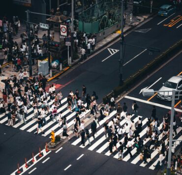 Soledad hiperconectada: Fotografía cenital de alto contraste sobre un cruce peatonal. Una marea humana de cientos de transeúntes, reducidos a siluetas desde las alturas, atraviesa de forma asincrónica las gruesas franjas blancas de un paso de cebra. La composición resalta el asfalto gris oscuro, vehículos de colores sobrios detenidos y la señalética vial, creando una retícula mecánica donde el individuo se disuelve en un flujo de movimiento masivo y geométrico.