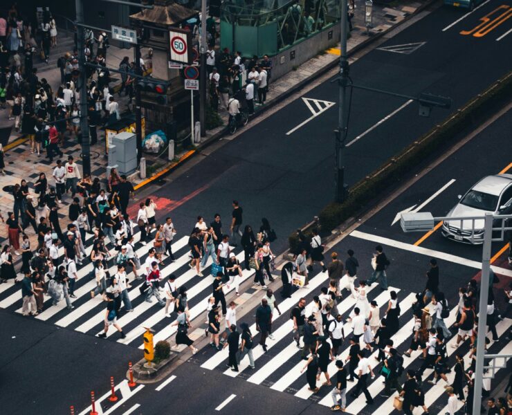 Soledad hiperconectada: Fotografía cenital de alto contraste sobre un cruce peatonal. Una marea humana de cientos de transeúntes, reducidos a siluetas desde las alturas, atraviesa de forma asincrónica las gruesas franjas blancas de un paso de cebra. La composición resalta el asfalto gris oscuro, vehículos de colores sobrios detenidos y la señalética vial, creando una retícula mecánica donde el individuo se disuelve en un flujo de movimiento masivo y geométrico.