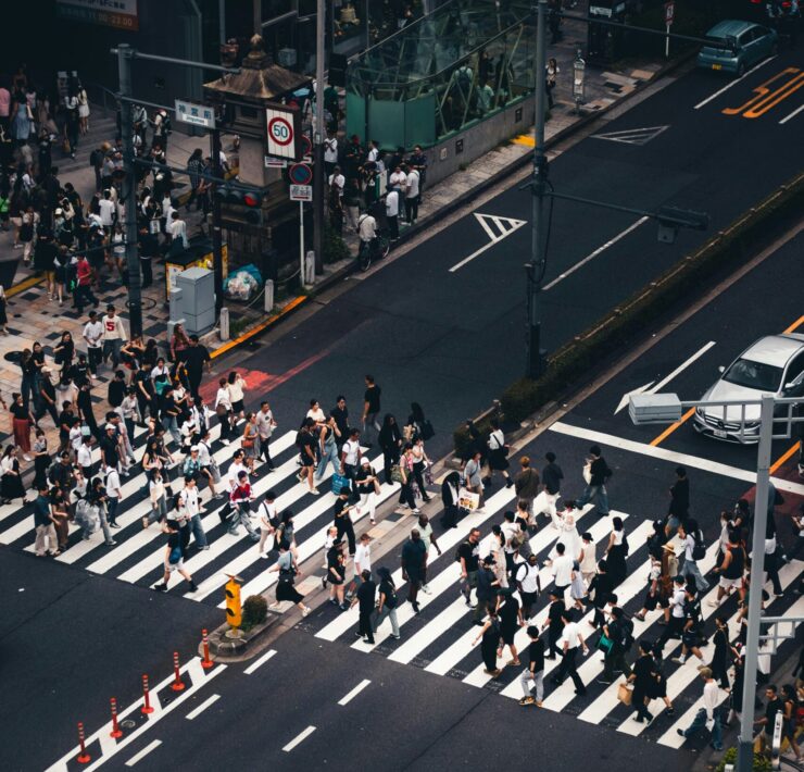 Soledad hiperconectada: Fotografía cenital de alto contraste sobre un cruce peatonal. Una marea humana de cientos de transeúntes, reducidos a siluetas desde las alturas, atraviesa de forma asincrónica las gruesas franjas blancas de un paso de cebra. La composición resalta el asfalto gris oscuro, vehículos de colores sobrios detenidos y la señalética vial, creando una retícula mecánica donde el individuo se disuelve en un flujo de movimiento masivo y geométrico.