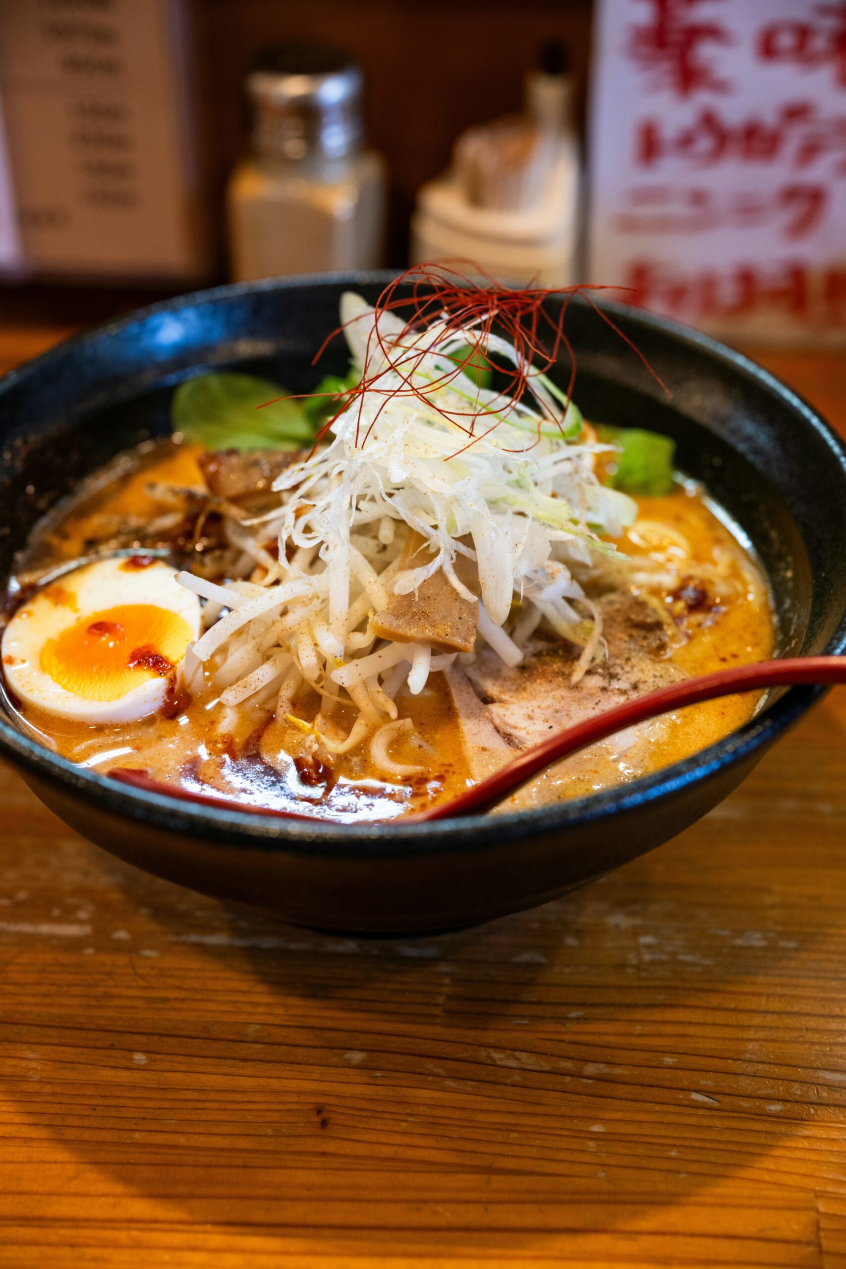 Ramen en Tokio: Foto en primer plano de un bowl de ramen tonkotsu. El caldo es de color crema intenso, cubierto por fideos, la mitad de un huevo marinado con la yema líquida, una rodaja de carne de cerdo y una montaña de cebolleta finamente picada. Unas hebras rojas de chile decoran la cima del plato. Se ve la punta de una cuchara de madera roja sumergida en el caldo.