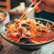Ramen en Tokio: Foto en primer plano de un bowl cerámico oscuro con ramen humeante. Se observa un caldo rojo vibrante con fideos finos, brotes de soja, mejillones y tentáculos de pulpo. Unos palillos de madera sostienen una porción de fideos elevándolos sobre el cuenco.