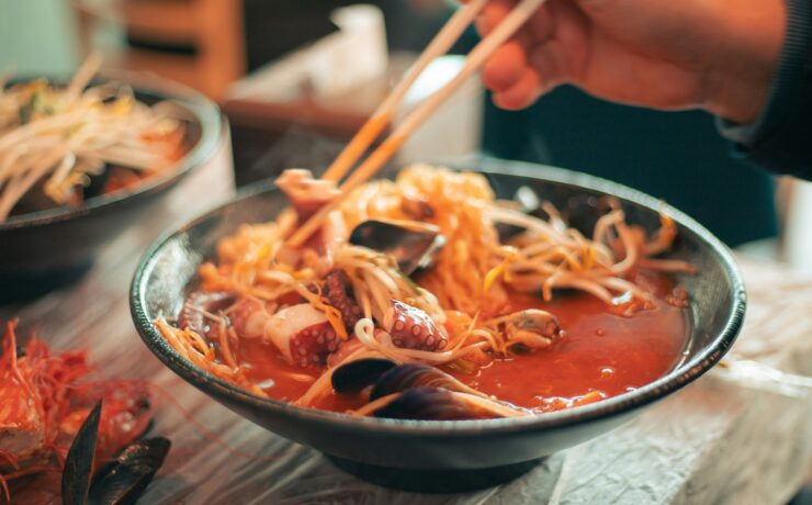 Ramen en Tokio: Foto en primer plano de un bowl cerámico oscuro con ramen humeante. Se observa un caldo rojo vibrante con fideos finos, brotes de soja, mejillones y tentáculos de pulpo. Unos palillos de madera sostienen una porción de fideos elevándolos sobre el cuenco.