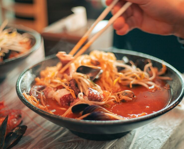 Ramen en Tokio: Foto en primer plano de un bowl cerámico oscuro con ramen humeante. Se observa un caldo rojo vibrante con fideos finos, brotes de soja, mejillones y tentáculos de pulpo. Unos palillos de madera sostienen una porción de fideos elevándolos sobre el cuenco.