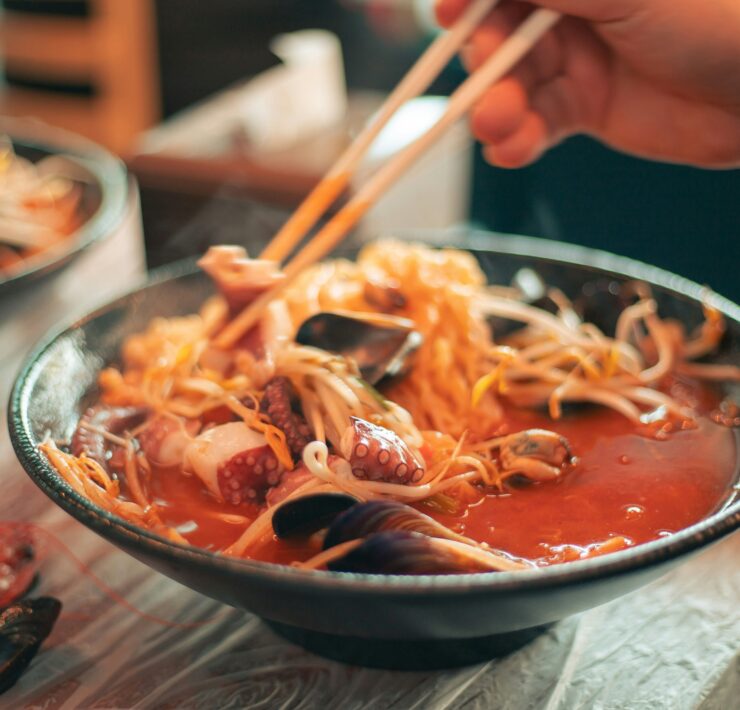 Ramen en Tokio: Foto en primer plano de un bowl cerámico oscuro con ramen humeante. Se observa un caldo rojo vibrante con fideos finos, brotes de soja, mejillones y tentáculos de pulpo. Unos palillos de madera sostienen una porción de fideos elevándolos sobre el cuenco.