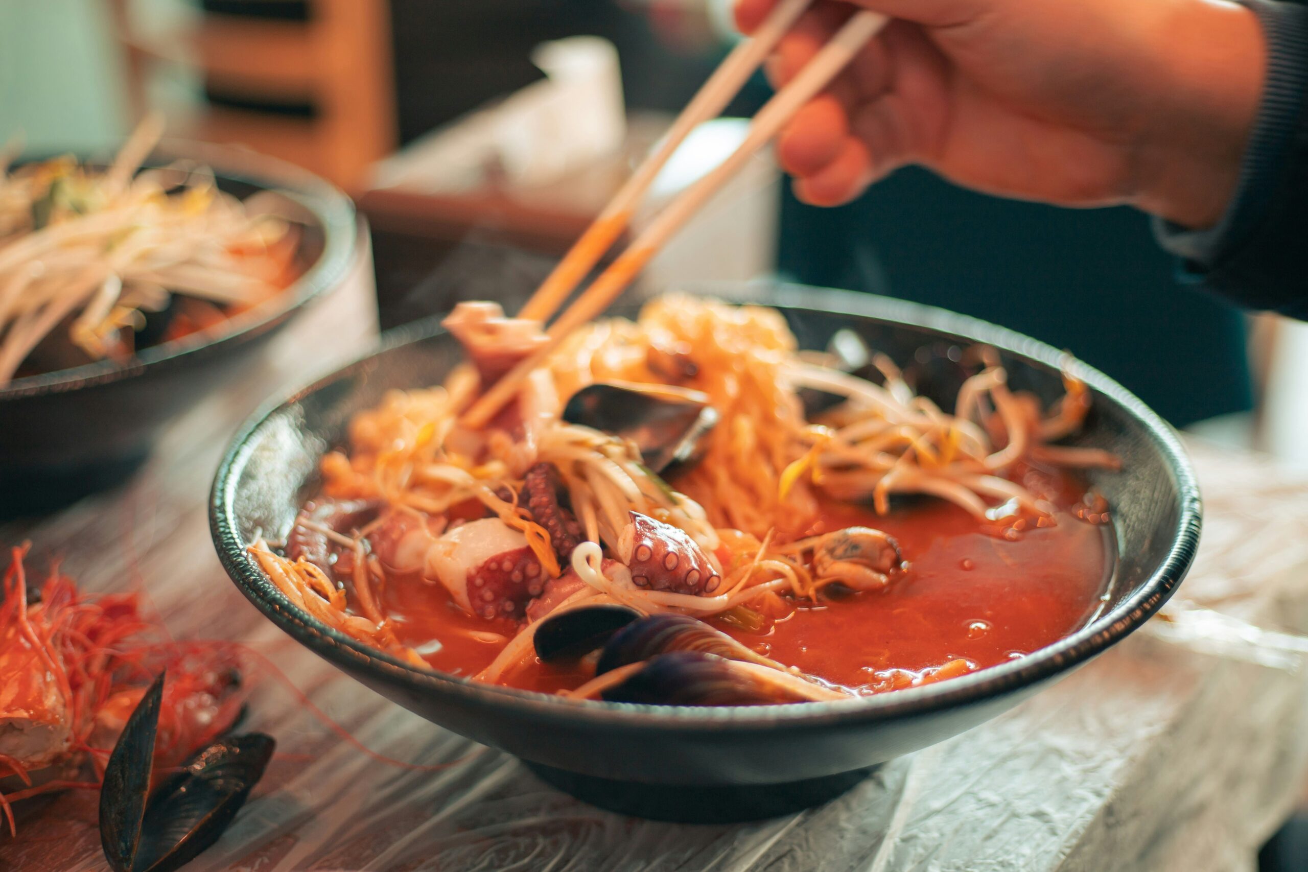 Ramen en Tokio: Foto en primer plano de un bowl cerámico oscuro con ramen humeante. Se observa un caldo rojo vibrante con fideos finos, brotes de soja, mejillones y tentáculos de pulpo. Unos palillos de madera sostienen una porción de fideos elevándolos sobre el cuenco.