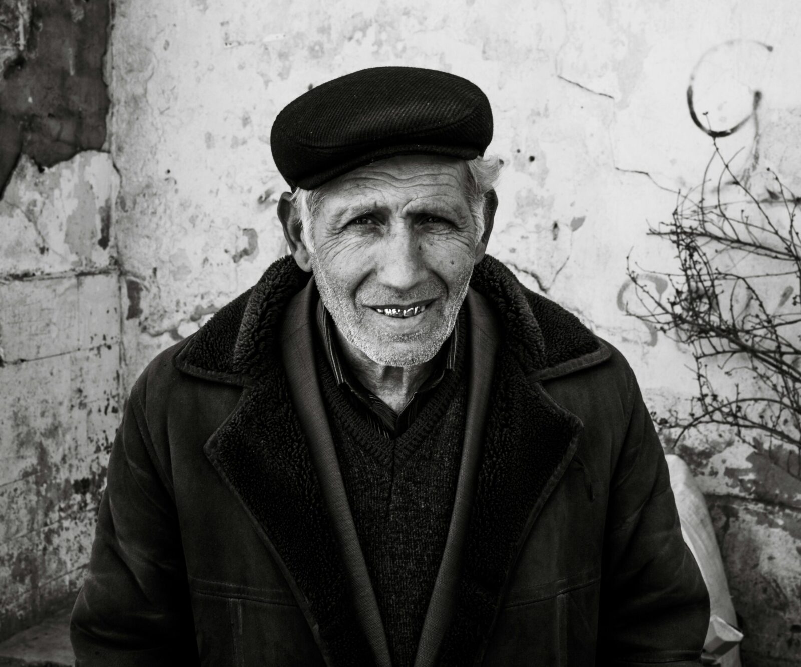 Retrato en primer plano de un hombre mayor con piel surcada de arrugas profundas y una sonrisa cálida que muestra sus dientes. Viste una gorra de paño oscuro, una chaqueta gruesa con cuello de textura de lana y una camisa a rayas bajo un jersey. El fondo es una pared blanca desgastada con grietas y una rama seca a la derecha. Una imagen que evoca la pérdida del padre a través de la serenidad y la vejez.