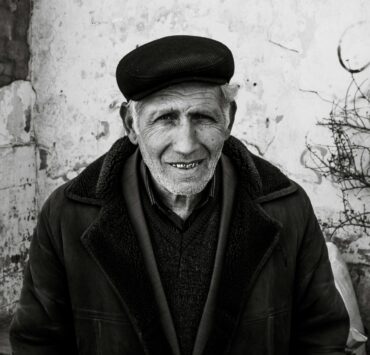 Retrato en primer plano de un hombre mayor con piel surcada de arrugas profundas y una sonrisa cálida que muestra sus dientes. Viste una gorra de paño oscuro, una chaqueta gruesa con cuello de textura de lana y una camisa a rayas bajo un jersey. El fondo es una pared blanca desgastada con grietas y una rama seca a la derecha. Una imagen que evoca la pérdida del padre a través de la serenidad y la vejez.
