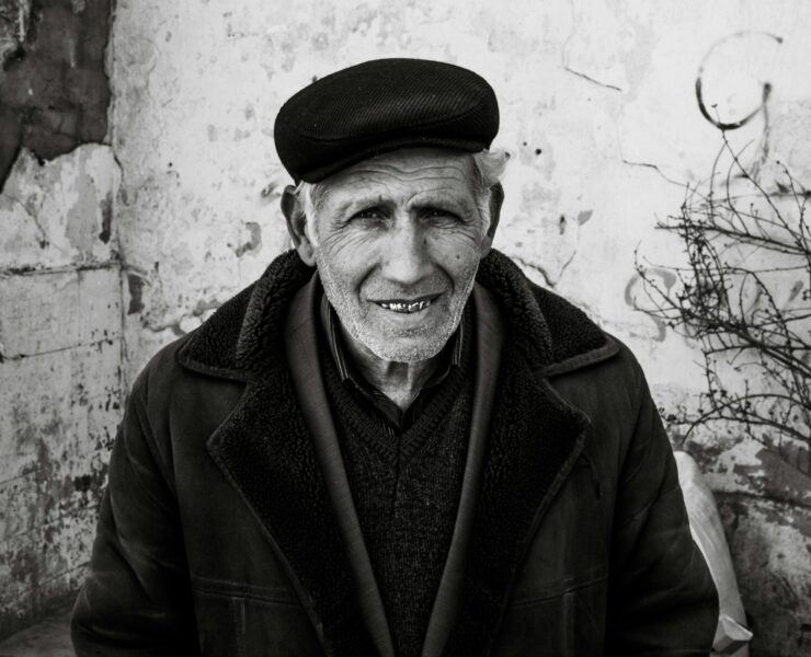 Retrato en primer plano de un hombre mayor con piel surcada de arrugas profundas y una sonrisa cálida que muestra sus dientes. Viste una gorra de paño oscuro, una chaqueta gruesa con cuello de textura de lana y una camisa a rayas bajo un jersey. El fondo es una pared blanca desgastada con grietas y una rama seca a la derecha. Una imagen que evoca la pérdida del padre a través de la serenidad y la vejez.