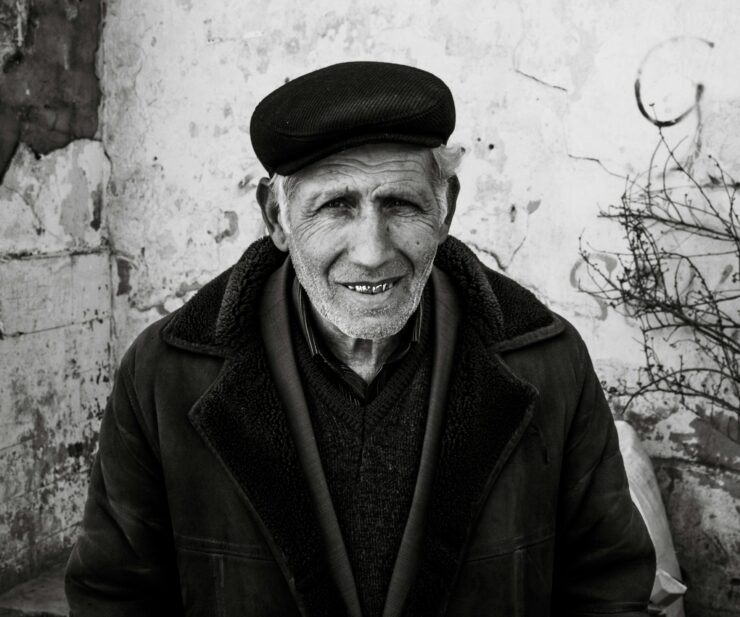 Retrato en primer plano de un hombre mayor con piel surcada de arrugas profundas y una sonrisa cálida que muestra sus dientes. Viste una gorra de paño oscuro, una chaqueta gruesa con cuello de textura de lana y una camisa a rayas bajo un jersey. El fondo es una pared blanca desgastada con grietas y una rama seca a la derecha. Una imagen que evoca la pérdida del padre a través de la serenidad y la vejez.