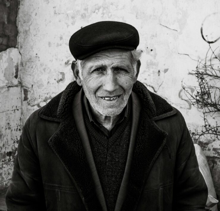 Retrato en primer plano de un hombre mayor con piel surcada de arrugas profundas y una sonrisa cálida que muestra sus dientes. Viste una gorra de paño oscuro, una chaqueta gruesa con cuello de textura de lana y una camisa a rayas bajo un jersey. El fondo es una pared blanca desgastada con grietas y una rama seca a la derecha. Una imagen que evoca la pérdida del padre a través de la serenidad y la vejez.