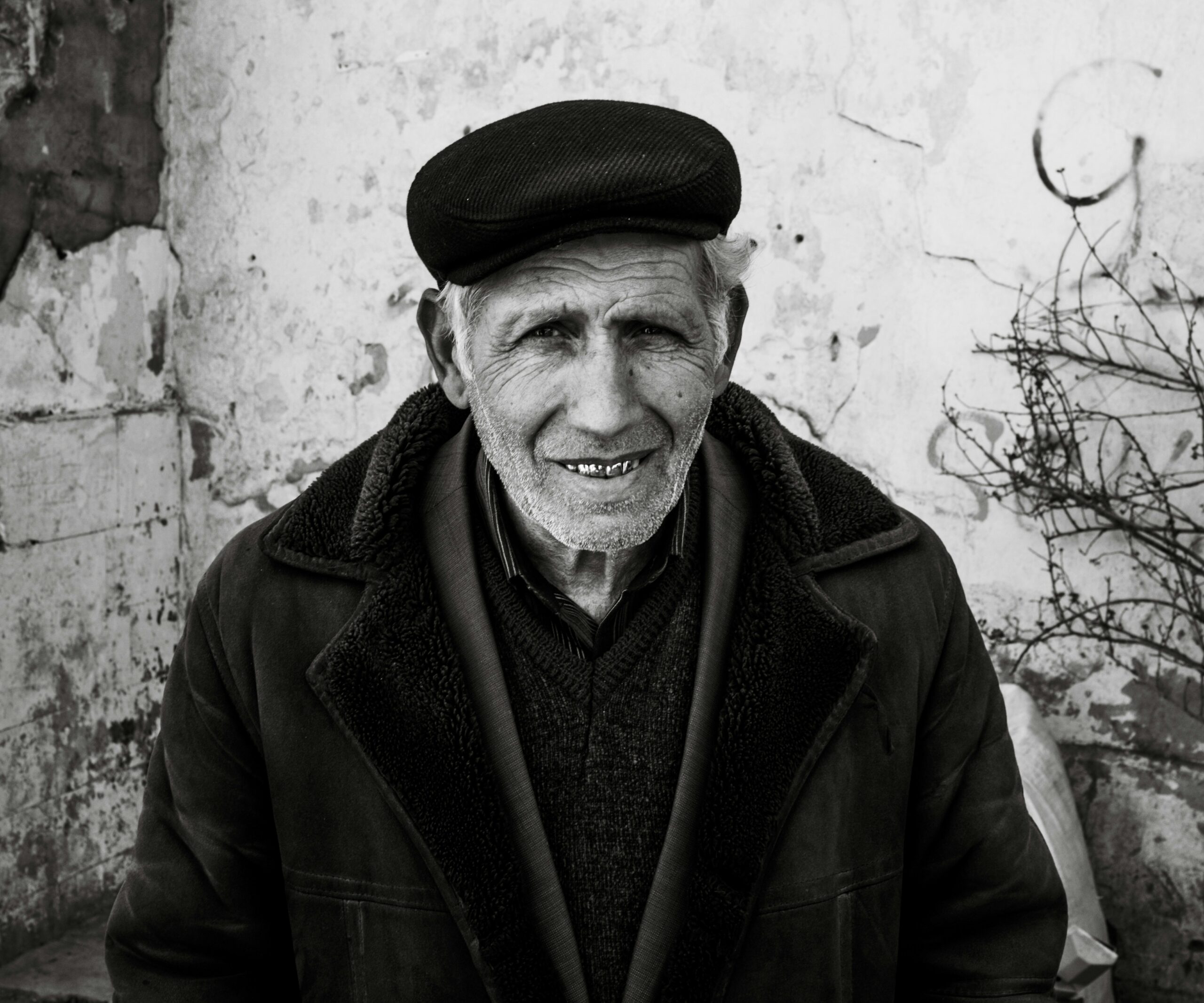 Retrato en primer plano de un hombre mayor con piel surcada de arrugas profundas y una sonrisa cálida que muestra sus dientes. Viste una gorra de paño oscuro, una chaqueta gruesa con cuello de textura de lana y una camisa a rayas bajo un jersey. El fondo es una pared blanca desgastada con grietas y una rama seca a la derecha. Una imagen que evoca la pérdida del padre a través de la serenidad y la vejez.