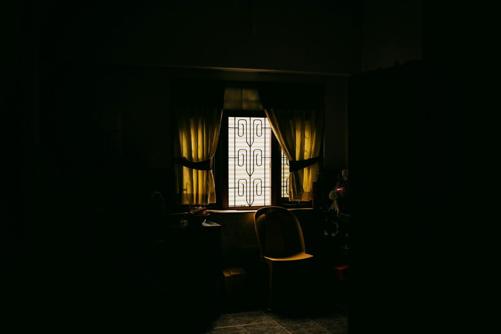 Fotografía de ambiente en plano medio que muestra el rincón de una cocina sumida en una penumbra profunda y lúgubre, donde la única fuente de luz proviene de una ventana con rejas de diseño rústico en el centro; la luz, filtrada y de un tono amarillento lánguido, ilumina una silla de plástico solitaria y una mesa desolada, simbolizando la ausencia, la invisibilidad y la soledad que vuelve polvo al ser.