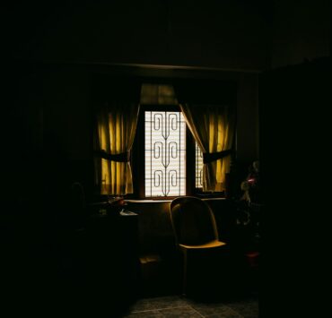 Fotografía de ambiente en plano medio que muestra el rincón de una cocina sumida en una penumbra profunda y lúgubre, donde la única fuente de luz proviene de una ventana con rejas de diseño rústico en el centro; la luz, filtrada y de un tono amarillento lánguido, ilumina una silla de plástico solitaria y una mesa desolada, simbolizando la ausencia, la invisibilidad y la soledad que vuelve polvo al ser.