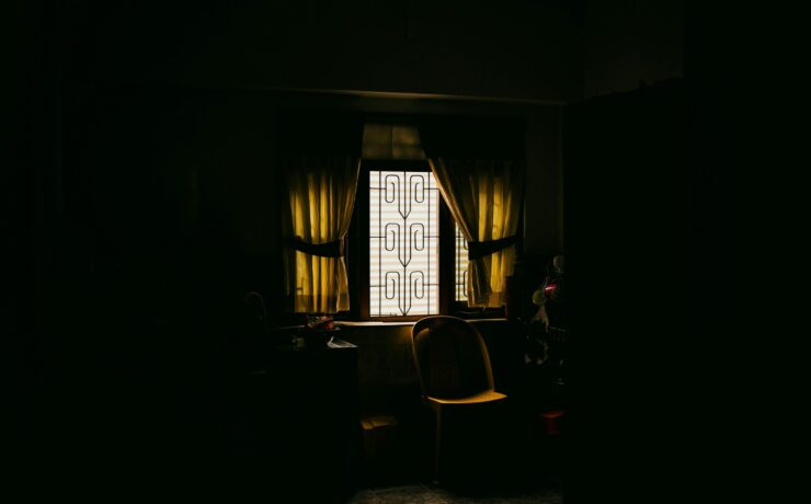 Fotografía de ambiente en plano medio que muestra el rincón de una cocina sumida en una penumbra profunda y lúgubre, donde la única fuente de luz proviene de una ventana con rejas de diseño rústico en el centro; la luz, filtrada y de un tono amarillento lánguido, ilumina una silla de plástico solitaria y una mesa desolada, simbolizando la ausencia, la invisibilidad y la soledad que vuelve polvo al ser.