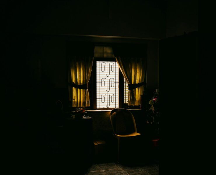 Fotografía de ambiente en plano medio que muestra el rincón de una cocina sumida en una penumbra profunda y lúgubre, donde la única fuente de luz proviene de una ventana con rejas de diseño rústico en el centro; la luz, filtrada y de un tono amarillento lánguido, ilumina una silla de plástico solitaria y una mesa desolada, simbolizando la ausencia, la invisibilidad y la soledad que vuelve polvo al ser.