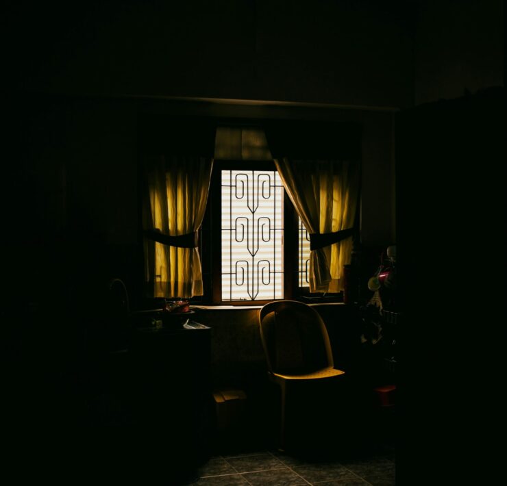 Fotografía de ambiente en plano medio que muestra el rincón de una cocina sumida en una penumbra profunda y lúgubre, donde la única fuente de luz proviene de una ventana con rejas de diseño rústico en el centro; la luz, filtrada y de un tono amarillento lánguido, ilumina una silla de plástico solitaria y una mesa desolada, simbolizando la ausencia, la invisibilidad y la soledad que vuelve polvo al ser.