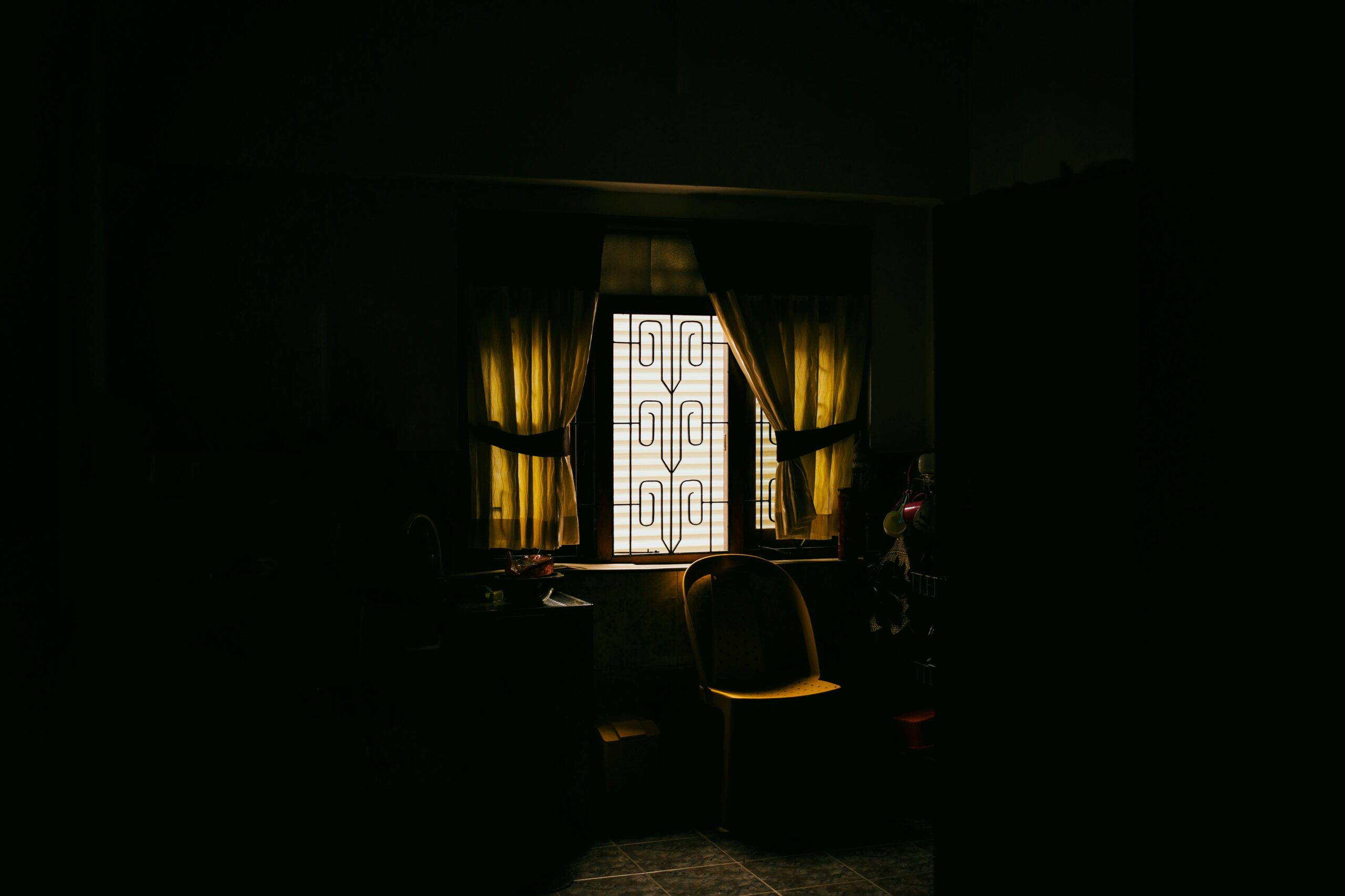 Fotografía de ambiente en plano medio que muestra el rincón de una cocina sumida en una penumbra profunda y lúgubre, donde la única fuente de luz proviene de una ventana con rejas de diseño rústico en el centro; la luz, filtrada y de un tono amarillento lánguido, ilumina una silla de plástico solitaria y una mesa desolada, simbolizando la ausencia, la invisibilidad y la soledad que vuelve polvo al ser.