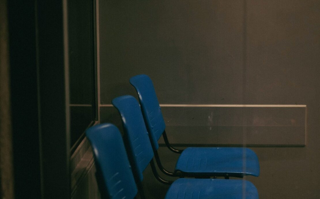 Vulnerabilidad humana: Imagen que muestra una fila de tres sillas de plástico azul en una sala de espera oscura y solitaria, vista a través de un cristal que refleja una atmósfera fría y burocrática.