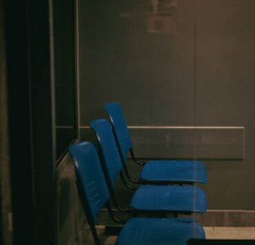 Vulnerabilidad humana: Imagen que muestra una fila de tres sillas de plástico azul en una sala de espera oscura y solitaria, vista a través de un cristal que refleja una atmósfera fría y burocrática.